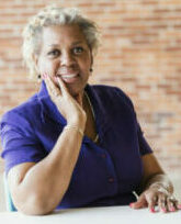 A mature African-American woman in her 50s sitting in an office, looking at the camera smiling.