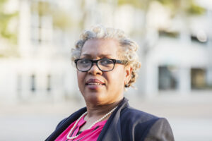 A mature, professional African-American businesswoman standing outdoors. She is in her 50s, well-dressed with short, gray hair and eyeglasses.