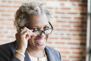 Close-up of the face of a mature African-American woman in her 50s with short gray hair. She is a businesswoman wearing a jacket and pearl necklace. She is smiling at the camera, looking over her eyeglasses.